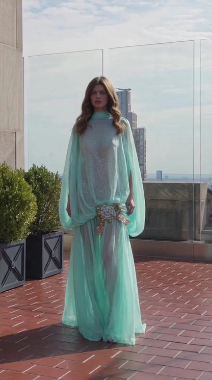 Front view of model wearing mint green high-neck cape sleeve maxi evening gown with floral brooch