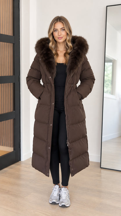 Front view of model wearing full-length brown hooded maxi puffer coat with faux fur trim