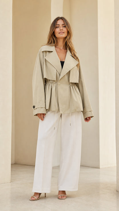 Full front view of model wearing light khaki short trench jacket with cinched drawstring waist and white wide-leg trousers