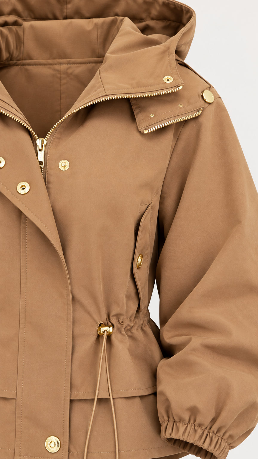 Close-up detail of camel utility jacket showing gold zipper, snaps, hood, and toggle drawstring
